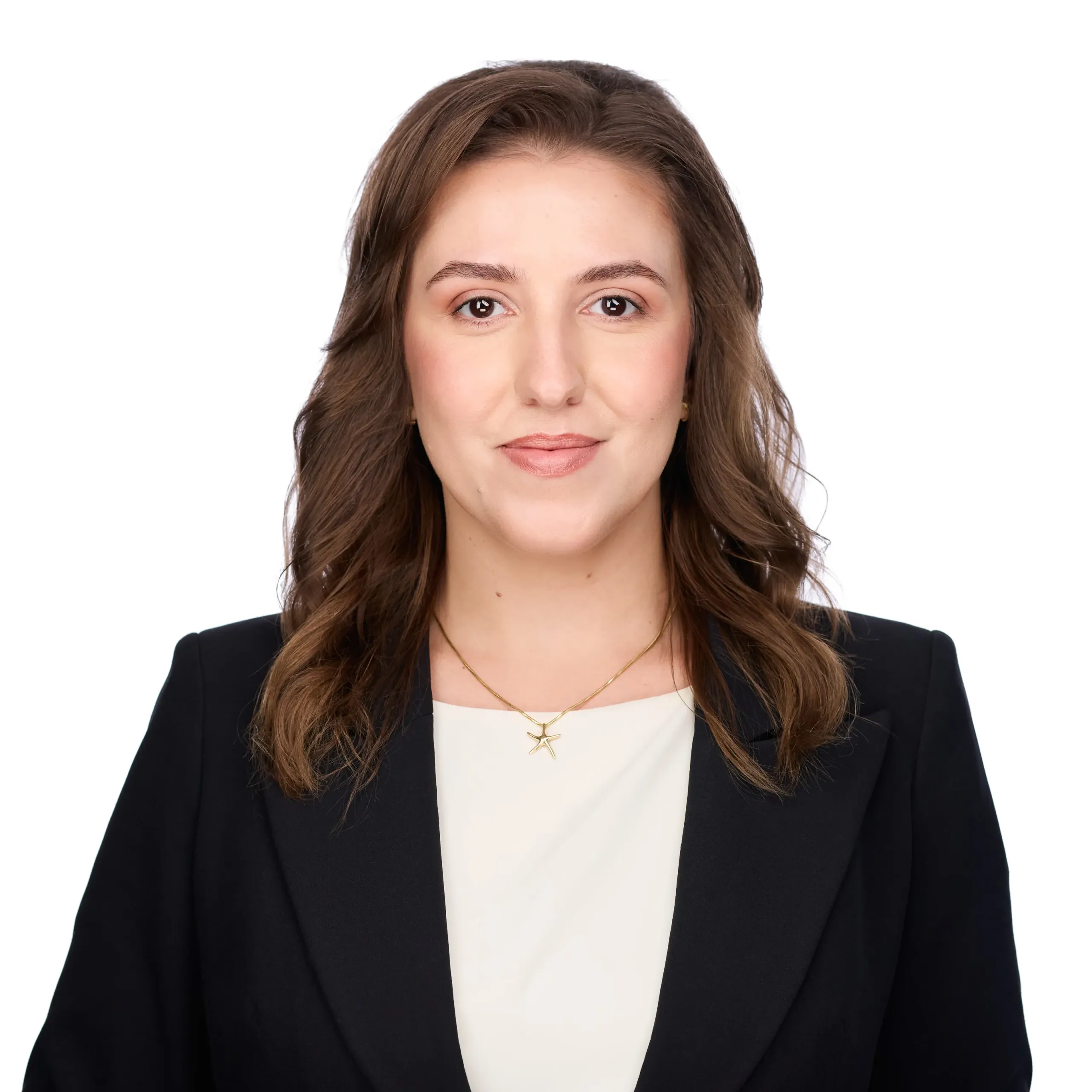 Woman with wavy brown hair in a black blazer over a cream top, smiling, wearing a gold necklace, on a white background.